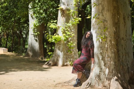 Mujer joven, guapa, alternativa y gordita, vestida con un vestido rojo, amándose y conectándose con la naturaleza apoyada en el tronco de un árbol grande. Millennial, regordeta, independiente, diferente.