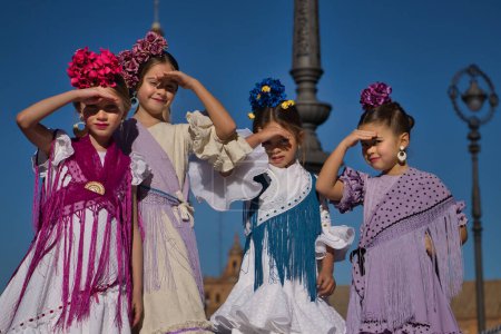 Four girls dancing flamenco, covering their eyes from the sun with their hands, in typical flamenco costumes on a bridge in a beautiful square in Seville. Dance concept, flamenco, typical Spanish.