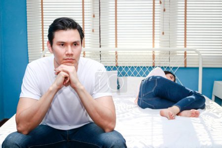 Téléchargez les photos : Family concept. Husband resents wife lying in bed He held his hands and feet on his chin, sat stressed at the end of the bed, feeling unhappy in the married life. couple problems - en image libre de droit