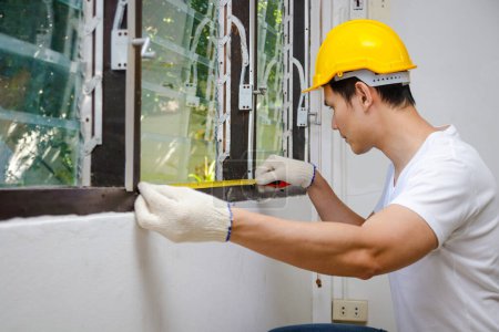 Photo for Asian handsome construction worker in yellow helmet Use meter tape to measure windows for home improvements. real estate concept residence. - Royalty Free Image