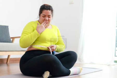 Fat asian woman exercising at home using a meter tape to measure her waist circumference. She looks shocked at the size. Sports concept, health care. weight loss