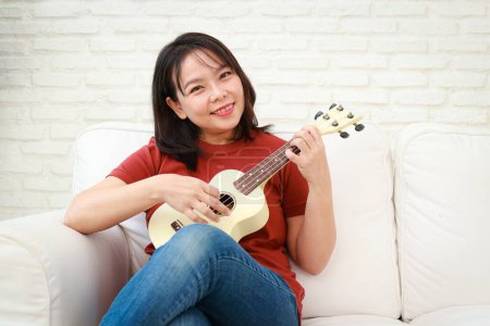 Woman playing music playing ukulele sitting at sofa in house. Music education concept. Artists, Bands and Live Performances