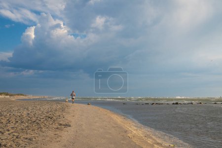 Man running with dog at sandy beach at morning, background overcast sky, Italy