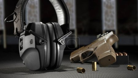 Foto de Pistola automática de 9 mm con munición y protección auditiva electrónica en una mesa con objetivos detrás - Imagen libre de derechos
