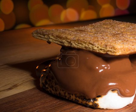 Smore on a wood table with chocolate melting and orange party lights glowing behind.