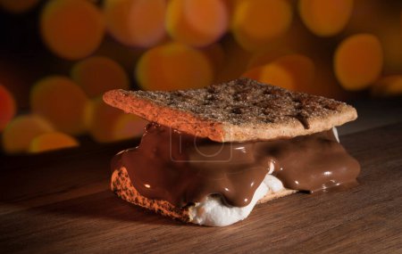 Huge smore with chocolate melting on the marshmallow on a wood table with orange party lights glowing behind.