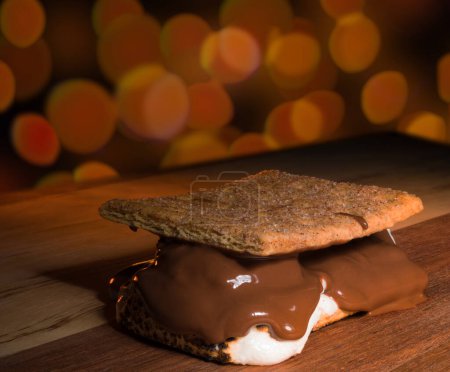 Tasty toasted s'more waiting on a wood table with orange light glowing brightly behind.