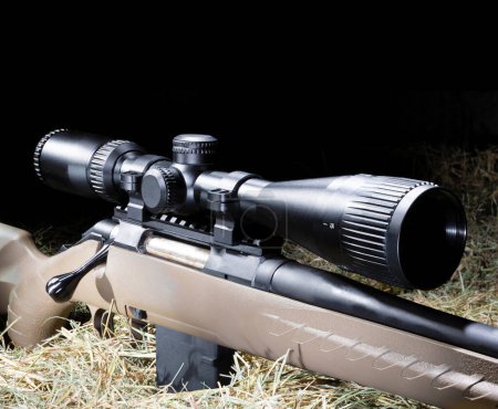 Rifle scope with adjustable magnification mounted on a bolt action rifle resting on a hay bale in a dark barn with copy space above. 