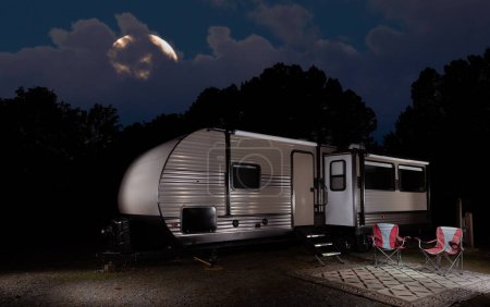 Travel trailer and camp all set up in a thick forest with the moon peeking through the clouds in the sky behind.