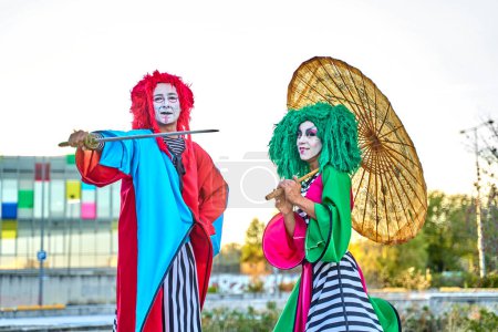 Photo for Full length of creative talented performers with face art in colorful costumes and wigs standing on stilts in park with Asian umbrella and sword during show, on sunny day - Royalty Free Image