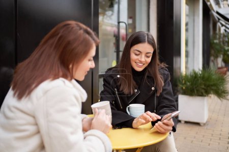 Foto de Female friends smiling while using a mobile phone sitting at coffee shop outdoors. Technology and friendship concept. - Imagen libre de derechos
