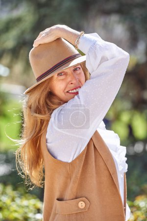 Photo for A beautiful blonde woman with long hair and a stylish hat looks directly at the camera with a warm smile, while surrounded by the lush greenery and natural beauty of a serene park setting - Royalty Free Image