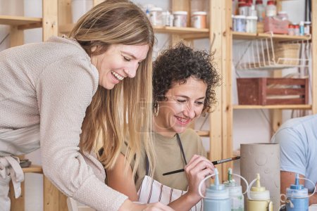 Foto de Dos mujeres alegres comparten una risa mientras pintan en una pieza de cerámica en un estudio de cerámica brillante y bien equipado - Imagen libre de derechos