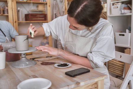 Foto de Artista atento usando una esponja para pintar una taza gris mientras gira sobre una rueda de cerámica en un estudio soleado - Imagen libre de derechos