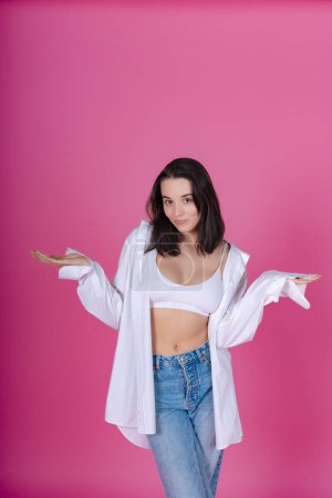 Photo for Confident woman with a playful expression balancing her open hands, wearing casual fashion on pink. - Royalty Free Image