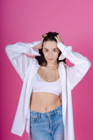 Photo for Confident young woman in a white bralette, open shirt, and blue jeans poses casually with a vibrant pink backdrop in a studio setting. - Royalty Free Image