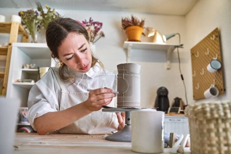 Focused artisan in white apron precisely detailing a clay vase on a spinning pottery wheel in studio