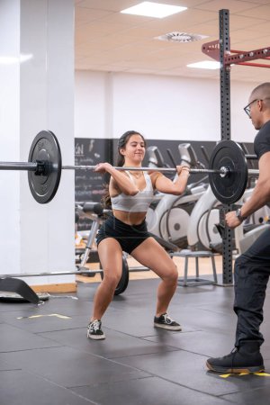 Fitness enthusiast performs a barbell squat in a gym under the watchful eye of her personal trainer, demonstrating proper form. 