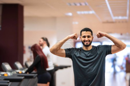 Hombre alegre mostrando los músculos con una flexión en el gimnasio, compañeros haciendo ejercicio detrás. 