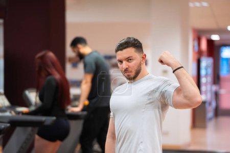 Muscular man in white tee flexes his bicep, with focused gym-goers behind him. 