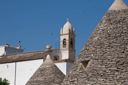 Foto de Campanario de la iglesia de Santa Lucía entre los tejados de los trulli de Alberobello, Italia. Trulli es una casa con un techo de piedra cónica que termina en un pináculo. - Imagen libre de derechos