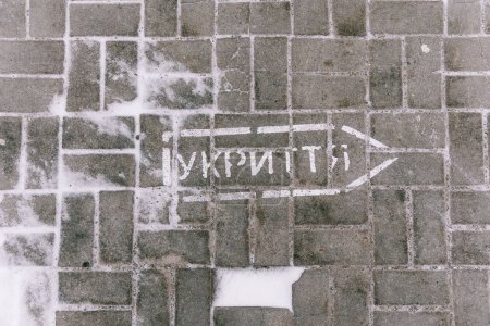Foto de Inscription in Ukrainian on the pavement tile "Shelter" with a drawing of an arrow indicating which direction to move in the event of the sound of an air-raid siren for evacuation - Imagen libre de derechos