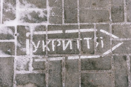 Inscription in Ukrainian on the pavement tile "Shelter" with a drawing of an arrow indicating which direction to move in the event of the sound of an air-raid siren for evacuation