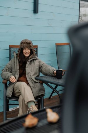 stilvolles Mädchen in warmer Jacke und Hut im Hof eines Touristenhauses grillt Fleisch und trinkt Kaffee, genießt einen ländlichen Urlaub und eine natürliche Landschaft