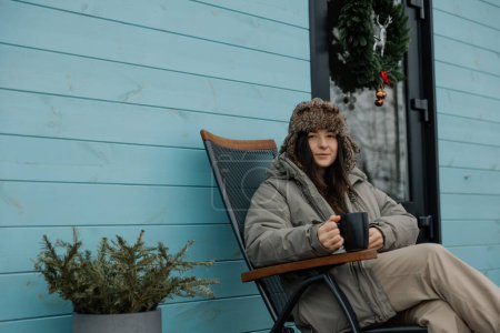 stilvolles Mädchen in warmer Jacke und Hut im Hof eines Touristenhauses grillt Fleisch und trinkt Kaffee, genießt einen ländlichen Urlaub und eine natürliche Landschaft