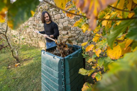 Téléchargez les photos : Un homme mélange les déchets organiques avec des feuilles sèches dans un bac à compost extérieur placé dans un jardin pour recycler les déchets domestiques et de jardin. Concept de recyclage et de durabilité - en image libre de droit