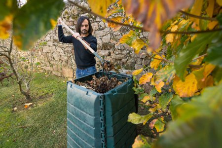 Foto de Un hombre está mezclando los desechos orgánicos con hojas secas en un cubo de compost al aire libre colocado en un jardín para reciclar los desechos domésticos y del jardín. Concepto de reciclaje y sostenibilidad - Imagen libre de derechos