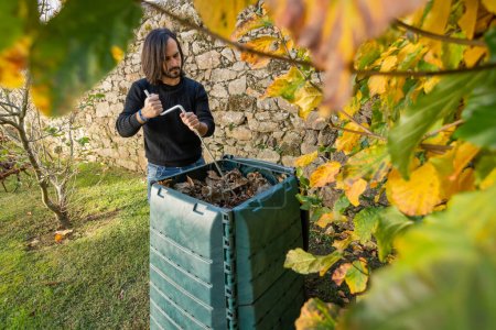 Un hombre está mezclando los desechos orgánicos con hojas secas en un cubo de compost al aire libre colocado en un jardín para reciclar los desechos domésticos y del jardín. Concepto de reciclaje y sostenibilidad