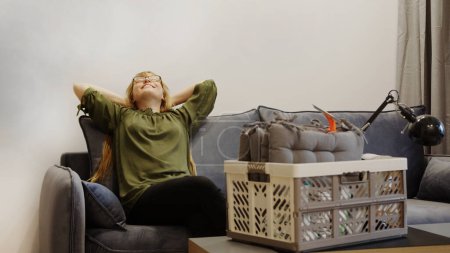 Photo for Happy woman resting on the couch after moving in and a box on the table, satisfied homeowner enjoying her first day in new house. High quality photo - Royalty Free Image
