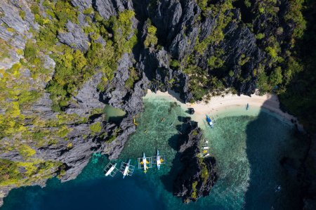 Luftaufnahme verschiedener touristischer Bangka-Boote an einem sonnigen Tag, mit seinem tropischen Strand und den wunderschönen felsigen Bergen, die Secret Lagoon, El Nido, Palawan umgeben.