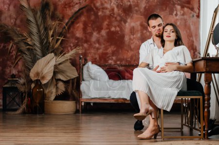 Photo for A pregnant girl and a man are sitting on a chair in an embrace in a brown room - Royalty Free Image