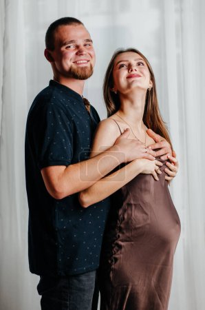 Photo for A pregnant girl in a brown dress stands in an embrace with a man - Royalty Free Image