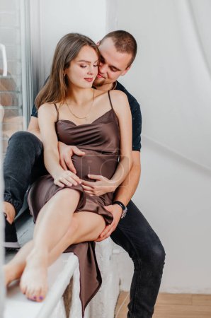 Photo for A man and a pregnant woman are sitting on a windowsill and hugging each other - Royalty Free Image