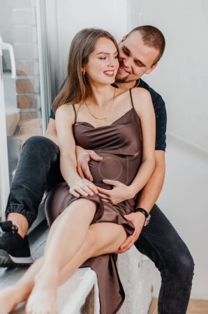 Photo for A man and a pregnant woman are sitting on a windowsill and hugging each other - Royalty Free Image