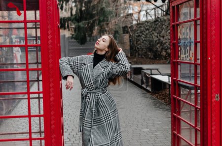 Photo for A girl in a gray coat stands by a red telephone booth - Royalty Free Image