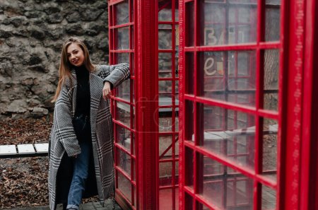 Photo for A girl in a gray coat stands by a red telephone booth - Royalty Free Image
