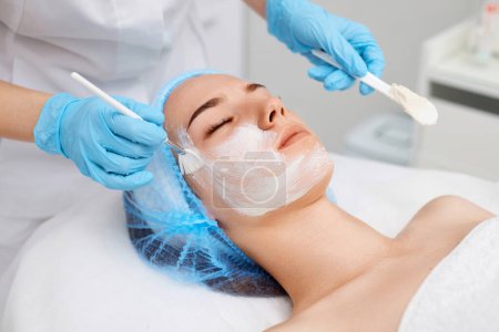 Photo for Beautician applies the mask to the face of beautiful young woman in the spa salon. Anti-aging, facial skin care concept. - Royalty Free Image