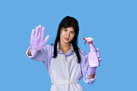 beautiful woman in rubber gloves and cleaner apron showing stop gesture isolated on blue background. focus on glove