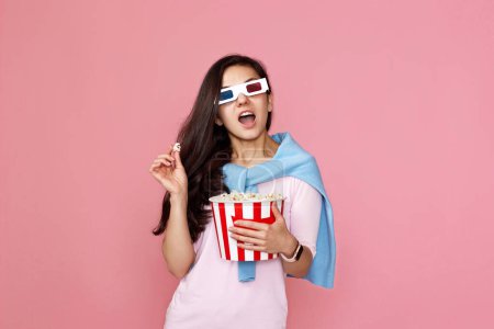 Foto de Mujer sorprendida viendo película de terror sosteniendo cubo de palomitas de maíz aislado sobre fondo rosa - Imagen libre de derechos