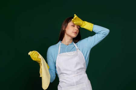 Photo for Tired woman in yellow gloves and cleaner apron on green background - Royalty Free Image