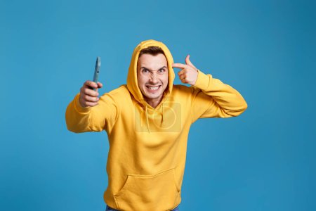 Foto de Hombre enojado y frustrado en sudadera amarilla sosteniendo el teléfono móvil y sentirse indignado aislado sobre fondo azul - Imagen libre de derechos