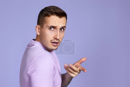 homme indigné en t-shirt pose une question et regarder en arrière-plan par-dessus son épaule sur fond violet. Qu'est ce que tu veux ? espace de copie