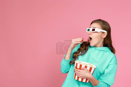 Foto de Asustada aterrorizada mujer morena rizada en sudadera viendo película de terror, sosteniendo cubo de palomitas de maíz sobre fondo rosa - Imagen libre de derechos
