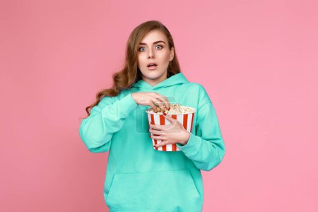 Foto de Terrified curly brunette girl in sweatshirt watching horror movie , holding bucket of popcorn on pink background - Imagen libre de derechos