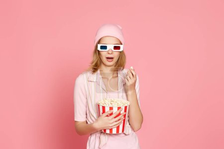 Foto de Amazed woman watching movie film, holding bucket of popcorn on pink background - Imagen libre de derechos