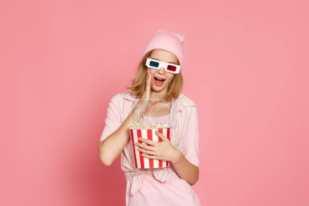 Foto de Amazed woman in 3d glasses holding bucket of popcorn on pink background - Imagen libre de derechos